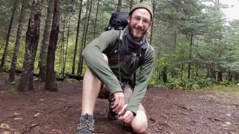 Alex Pretti kneels in front of the camera wearing hiking gear in a green forest.