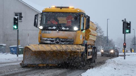 A yellow snow plough on a snow covered street with a car behind and traffic lights alongside.