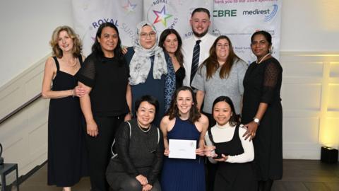 Ten people posing for a photo with their award. Seven people are standing up and three people crouching down. They are all smiling at the camera. 