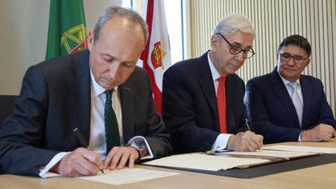Three men in suits signing documents. There are flags behind them.