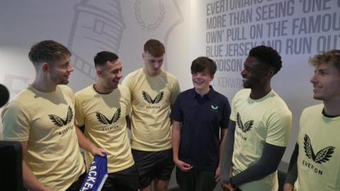 Mackenzie, in a dark blue polo-neck t-shirt, beams at the camera surrounded by five Everton players wearing yellow t-shirts