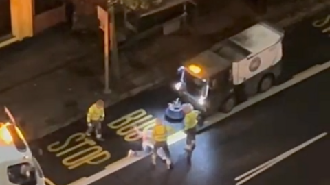 Three workers wearing hi-viz jackets stand around a man laying on his back on the street. Their faces are blurred. A lorry is parked on the left and a road sweeper is parked on the right.