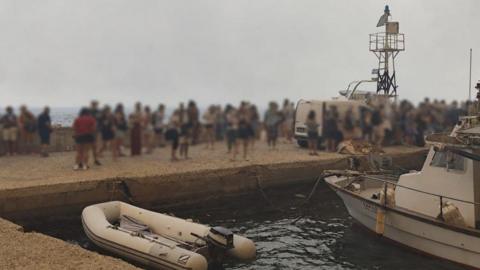 Blurred-out people stand on a marina pier while waiting to be evacuated in Agios Vasileios in Greece.