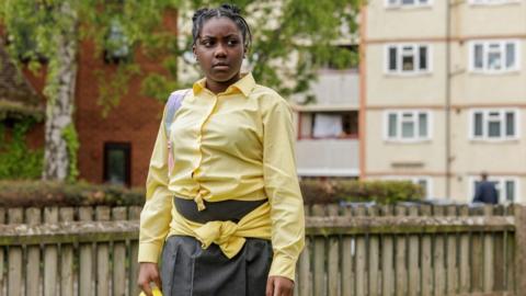 Girl with braided hair who is wearing a yellow shirt and grey skirt.