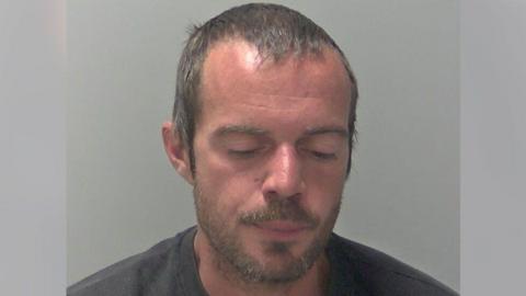 A man, Daniel Gibbs, looks down at the ground in a police mugshot. He has light brown, short hair and light brown stubble. He is wearing a dark-coloured top.