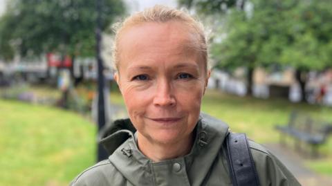 A woman in a green rain coat looks at the camera. She has pulled back blonde hair and a backpack hangs from her right shoulder. Behind her, greenspace in Heckmondwike is blurred.