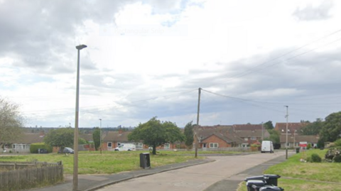 A street view of St Austell Road in Leicester, showing a grey road, lined by grass verges and houses