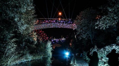 A canal bridge at night. The bridge is illuminated with purple and red circular lights. The trees along the canal are also illuminated with white light.