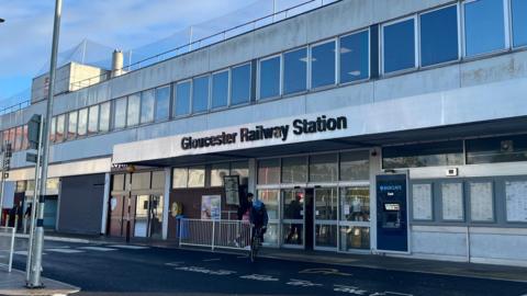 A cyclist rides away from the entrance of Gloucester Railway Station as another passenger is about to go inside. The building is grey and white, pictured on a sunny day with blue skies. There are notice boards and an ATM machine on the exterior wall of the building, to the right of the entrance doors. On the left side of the doors there is the train departures board.