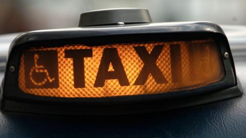 An illuminated taxi sign on top a vehicle. It has a disability sign, depicting a person in a wheelchair, to the left of the sign.