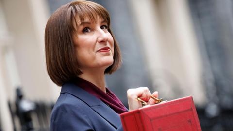 Rachel Reeves who has a brown bob haircut poses side-on holding up her red box. She is wearing a dark blue suit and a purple blouse