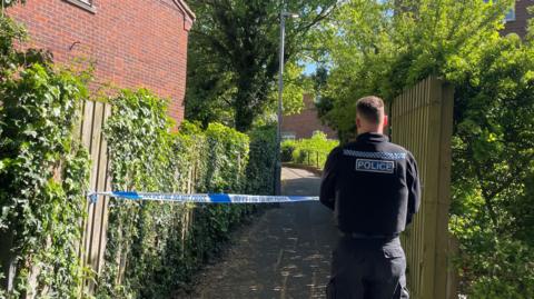 The back of a police officer in front of some blue police tape with a path behind the tape. The path goes between two fences with trees overhead.