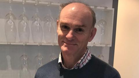 Philip Tuck in a blue jumper with a checked shirt, standing in front of shelves with wine glasses on.