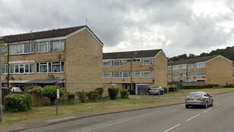 A large housing estate with four-two-story blocks. Cars can be seen on the road and in the distance. There are grass verges with shrubbery and trees in the background.