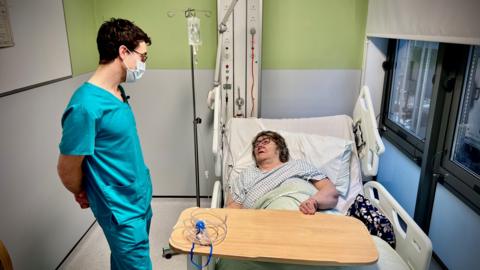 A doctor is standing in a side room in an A&E department.  He is talking to a 77-year-old woman who is lying in bed.  She is breathing oxygen through a tube that goes into her nose.  He is wearing a face mask.