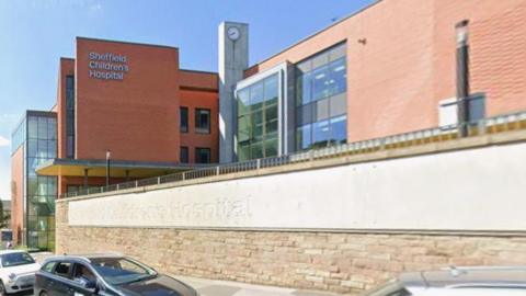 The exterior of a large, modern hospital building. The main structure is made of red brick with sections of glass and metal, giving it a contemporary appearance. On the left side of the building, there is a prominent sign that reads “Sheffield Children’s Hospital”.