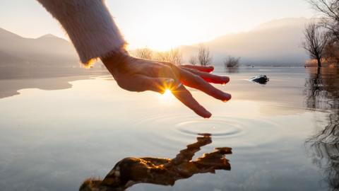 A hand touching and caressing water surface of beautiful lake at sunset, mountain view