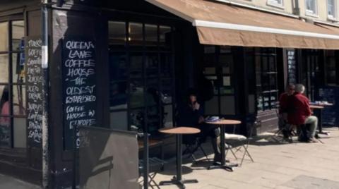 The sun is shining on tables and chairs outside a cafe. A faded brown canopy hangs over the front of the cafe. White chalk writing on a black sign outside the coffee house reads Queen's Lane Coffee House "the oldest established coffee house in Europe since 1654. An older couple sit at one of the tables at the far corner of the image