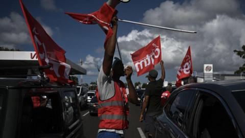 Men in gilets wave flags of the French union CGT Martinique during aa go slow operation on the ring road leading to the airport in Fort-de-France, in the French Caribbean island of Martinique.