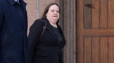 Esther Baker walking into court, she wears a black patterned dress under a black blazer. She has long brown hair.