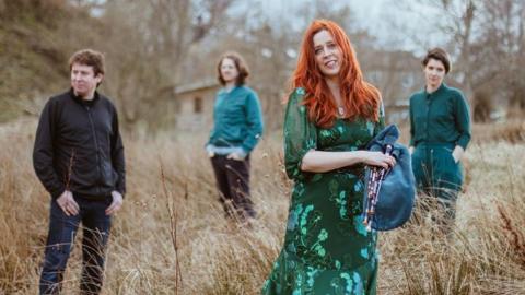 Four members of the band Kathryn Tickell and The Darkening are standing in a grassy field  with trees in the background 