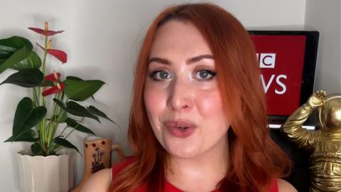 Alice Cruickshank looks at the camera, a plant, a screen with BBC News and a gold astronaut sit behind her