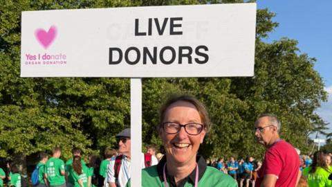 Jo Jones holds a 'Live Donors' sign at the British Transplant Games in Oxford. She is wearing glasses and a green polo shirt. Dozens of people are stood behind her. She has a broad smile on her face.
