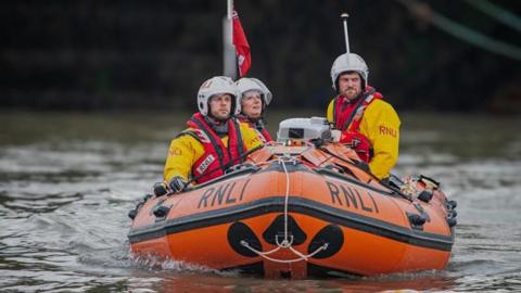 Three RNLI rescue crew members in yellow jackets and helmets are in an orange inflatable lifeboat on calm water. The boat is marked "RNLI" and flies a red maritime flag. They look alert and focused, possibly on a mission or training.