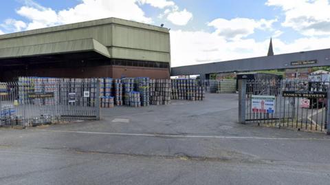 The entrance to a brewery site - a building is on the left of the frame with barrels piled up next to it. Black signs that say "welcome to Banks's Brewery" can be seen on the fences either side of the entrance.                