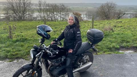 A woman with long grey and black hair is sitting on a motorbike wearing a black jacket and trousers. Behind her is a beautiful view of the Somerset levels. 