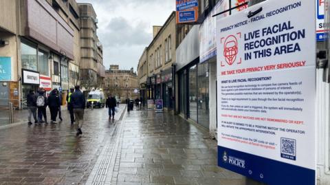 Close-up of a poster on a lamppost detailing the use of live facial recognition technology. In the background people walk past along a wet street lined with shops. A live recognition van can bee seen in the distance.