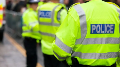 A shot from behind a police officer who is wearing a high-visibility jacket with the word POLICE written on it. Their face is not in the shot. There are two other officers, dressed in the same uniform, at the back of the photograph. 