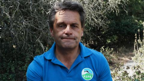 A head-and-shoulders image of a man standing in front of burnt hedging. He has a look of resignation. He is looking straight at the camera. 