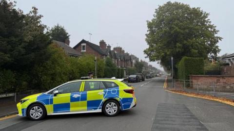 A police car parked sideways on a road leading to a residential street. A police officer can be seen standing on the other side of the car. Terraced houses line the left of the street in the distance with cars parked at the side of the road.