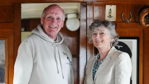 Henry Montgomery and Arabella Stuart-Smith are standing inside a caravan which has wooden stained walls that has photos and objects hanging up. Henry is wearing a white jumper which has a logo. He has short white hair and is smiling. Stuart-Smith has short grey hair. She is wearing a beige blazer and blue floral shirt, and is also smiling.