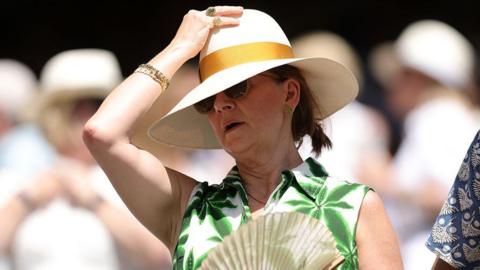 Woman wears hat and carries fan at Wimbledon in hot weather