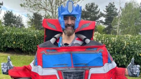 Matt Batchelor, with two arms held out, dressed in a costume, as Optimus Prime. He has a beard and is standing on grass with bushes behind him.