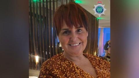 A close-up of a woman with short red hair wearing a gold glittery top smiles at the camera showing her teeth. Her face, neck, and shoulders are visible. She is sat in a room with ceiling to floor windows and white curtains in the background, in what appears to be a restaurant or bar. 