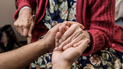 A pair of hands holding an elderly woman's hands. The woman wears a red cardigan and floral dress. She is seated.