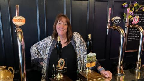 Hazel Smith stands behind the bar set up in a tap room created in the couple's front room. There are tap handles customised with brewery branding and there is a board with writing on to one side, decorated with flowers. Mrs Smith is wearing a black top and a patterned cardigan. She is smiling, with one hand on the bar, and has shoulder-length hair.