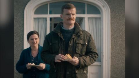 A woman looks in disgust at a moustached man who is in front of her. He is holding a plate of sandwiches