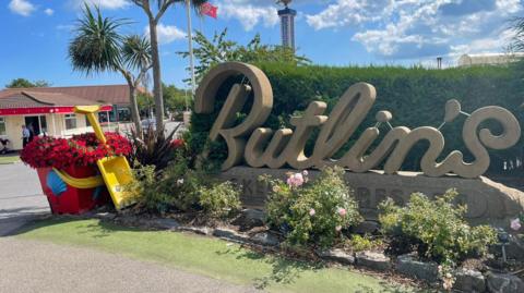 Entrance to the Butlin's holiday resort in Ingoldmells, Skegness. There is a plinth with the company's name on it and a giant yellow and red bucket and spade to the side. There is also a palm tree and a tower in the background.