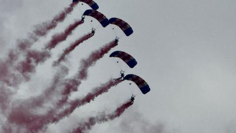 Five people with parachutes deployed falling from a grey sky as red-coloured smoke trails behind each of them.