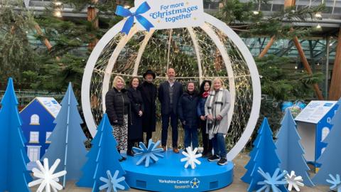 A large, open-front bauble in the Sheffield Winter Gardens, an indoor enclosure filled with trees and plants. It's stood on a blue, Yorkshire Cancer Research branded podium, and surrounded by blue cut-outs of Christmas Trees. Two information boards are positioned either side. Inside the bauble stand a group of representatives from the charity.