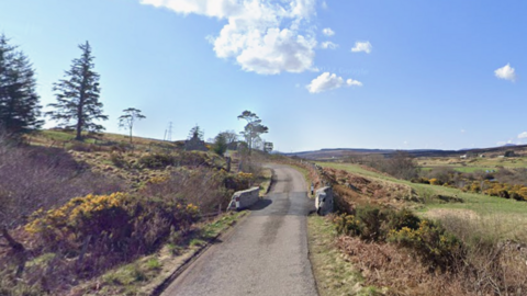 A single-track road and a small bridge with low stone walls. The surrounding landscape is of green fields and areas of gorse.