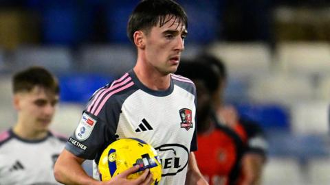 Exeter City's Tom Dean, wearing the club's white away kit with grey and pink trim, holds a ball in his right arm