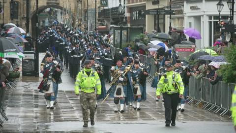 Police officers in high-visibility yellow jackets lead a pipe band with blue tartan kilts followed by RAF personnel in blue dress uniforms carrying rifles in a parade along Lincoln High Street past crowds of spectators
