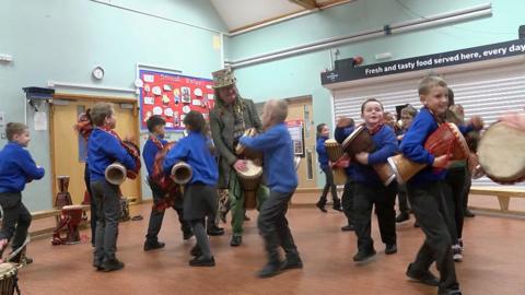 Pop star Tom Morley wears a green top hat with a long green coat. He is standing and banging a drum. He is surrounded by primary school children with drugs. They are in a school hall.