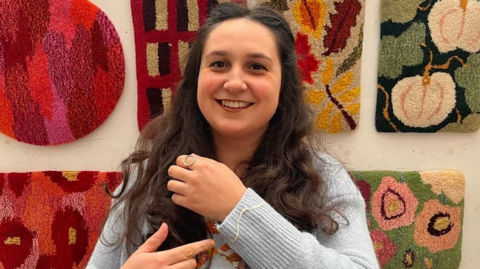 Jessie is in a mid-shot, smiling to the camera and pointing to her left arm, showing off the embroidery she added to the cuff of her grey cardigan. She has long, wavy dark hair, and there are several brightly coloured rugs she has made on display on the wall behind her
