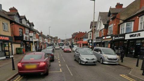 Evington Road with lots of cars on it, there are shops on either side of the road.